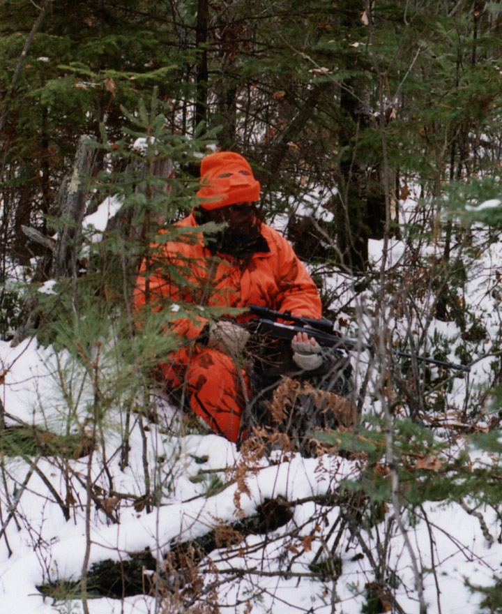 John, leaning against a tree stump, surrounded by small pines, wearing a camo face mask.